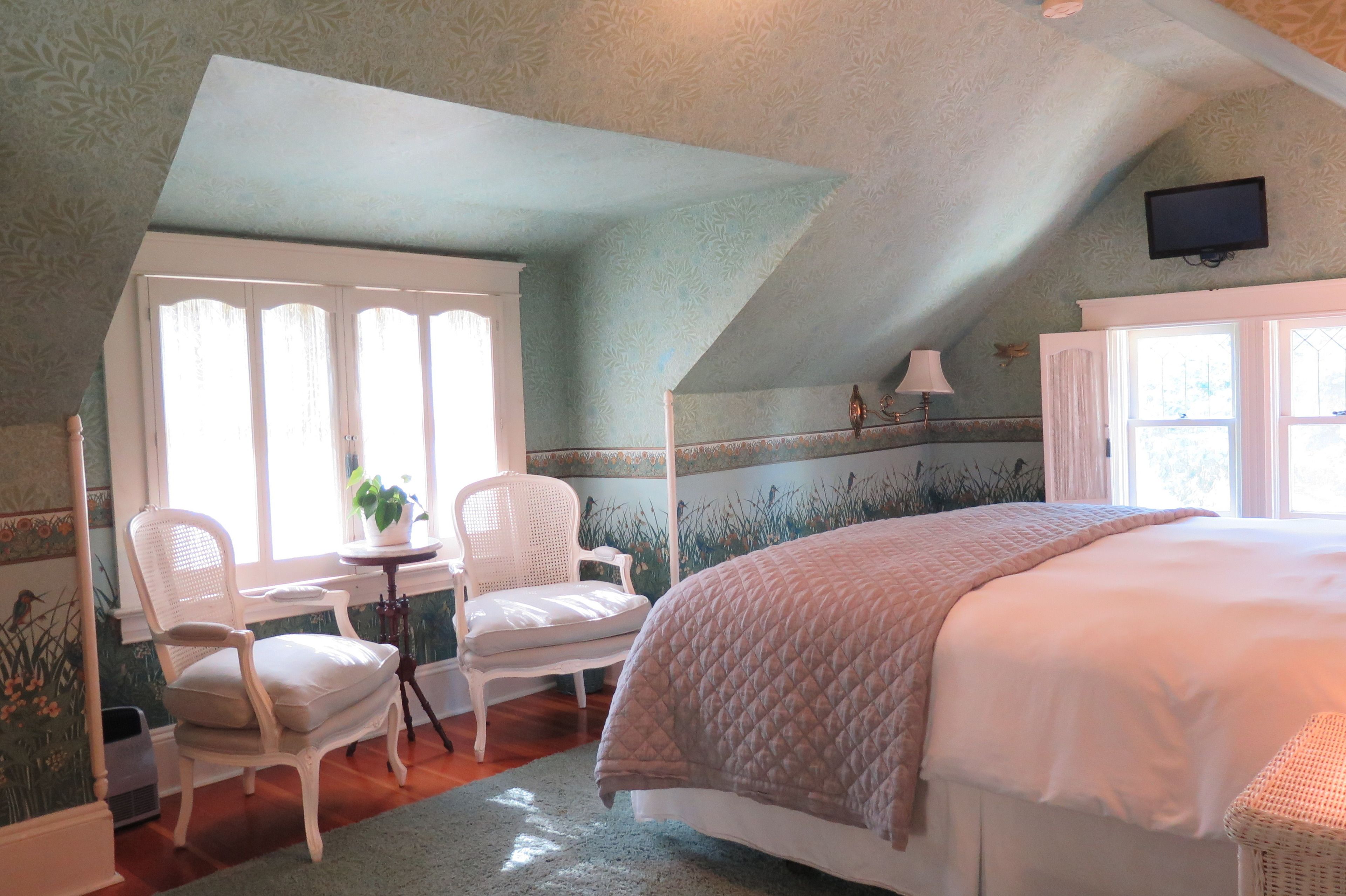 Een slaapkamer met een groot bed, een raam met gordijnen, een witte stoel en een tafeltje met een plant.