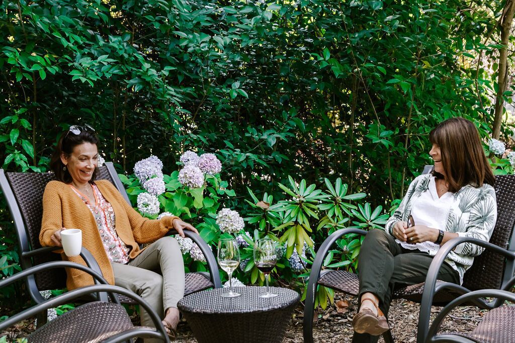 Two people seated on wicker chairs in a garden with lush greenery and flowering plants.