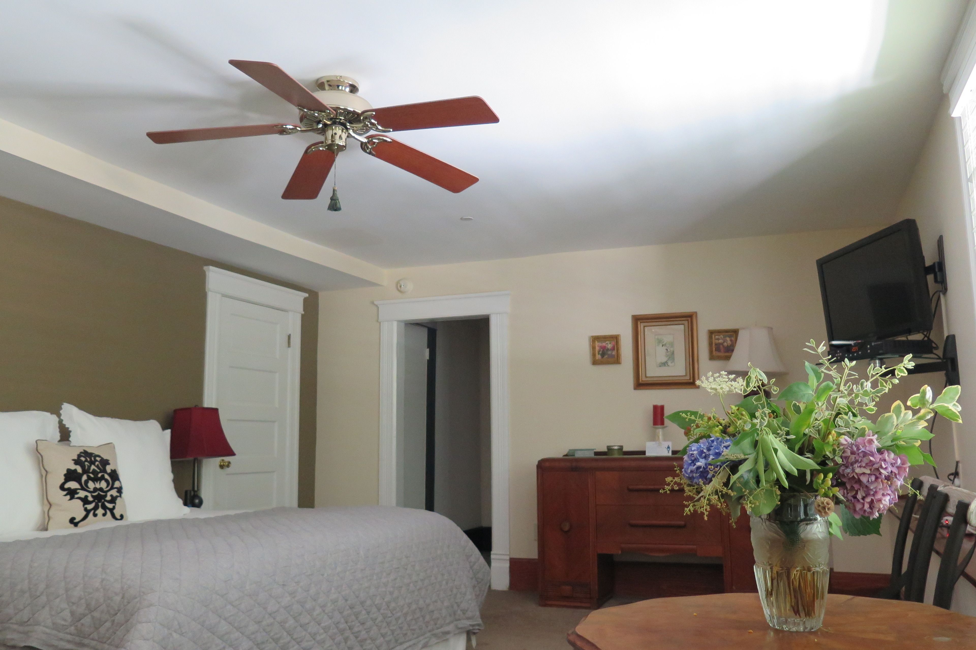 Una habitación de hotel con una cama, un escritorio, una silla, un florero con flores, un ventilador de techo y un televisor.