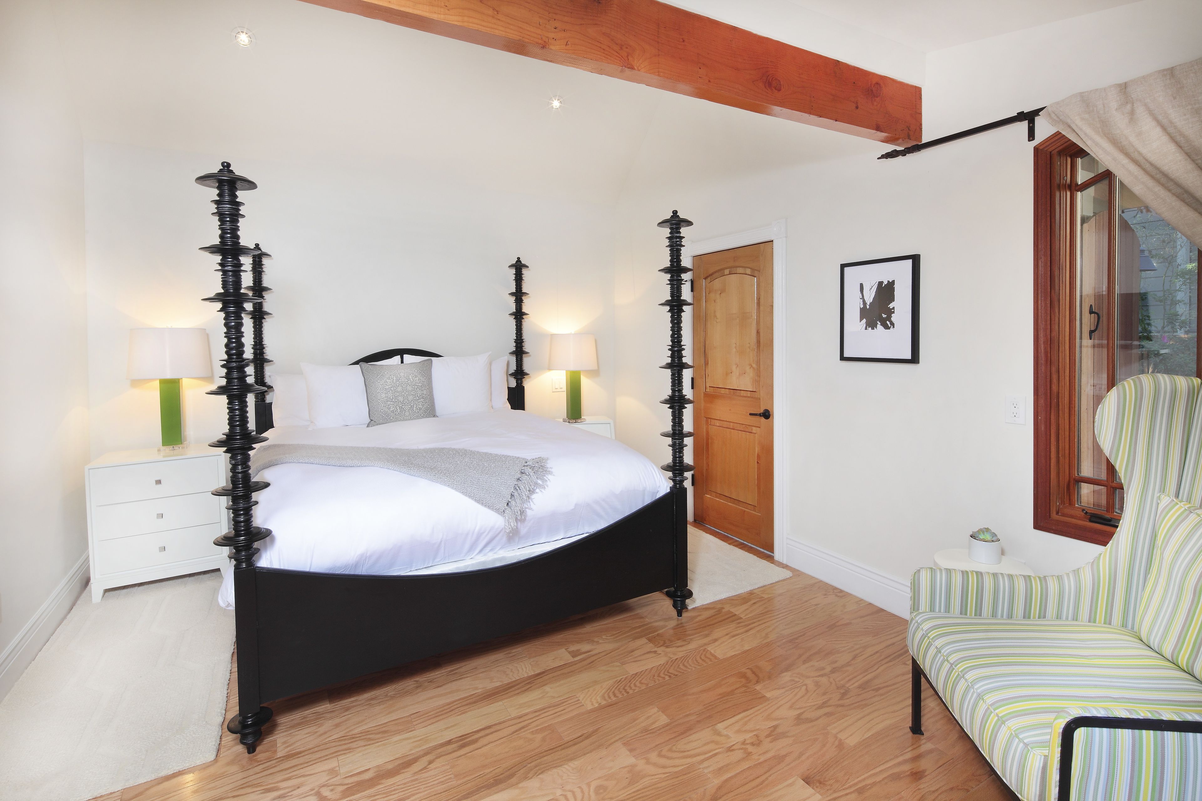 A bedroom with a four-poster bed, a wooden floor, a white nightstand, a green lamp, a striped armchair, and a wooden door.