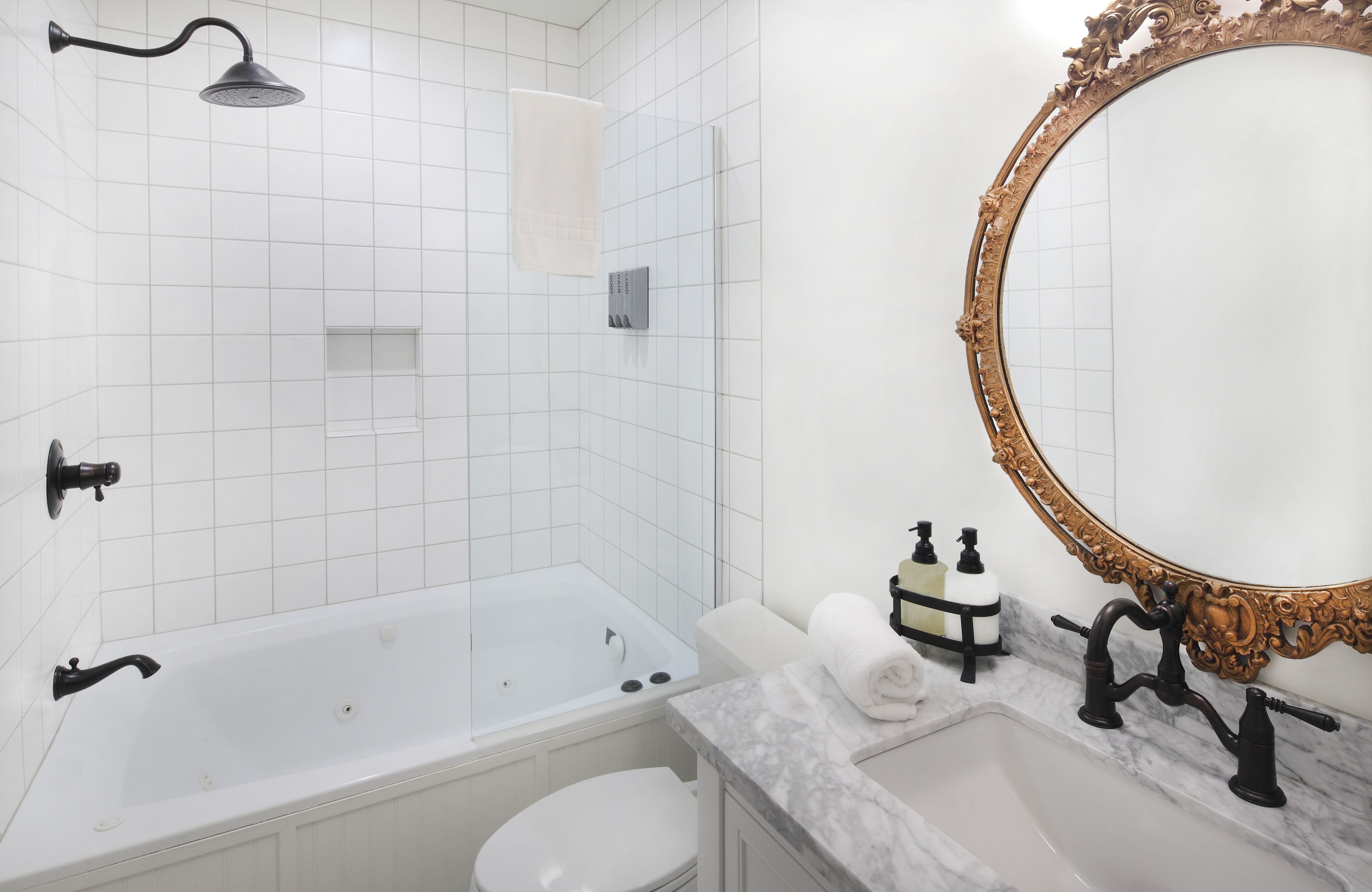 A bathroom with a white bathtub, a toilet, a sink with black faucets, a showerhead, and a large ornate mirror.