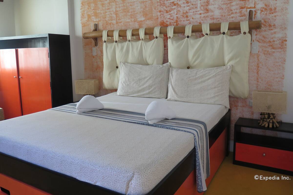 Una cama bien hecha con una colcha a rayas, dos almohadas y un cabecero con cortinas. Se aprecia una mesita de noche con una lámpara y un pequeño adorno. Un armario rojo es visible a la izquierda.