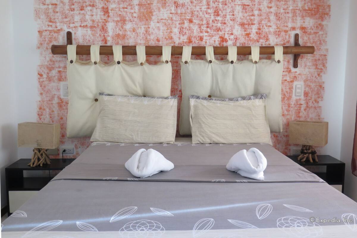 Una habitación de hotel con cama, dos almohadas, cabecera, mesitas de noche y lámparas.