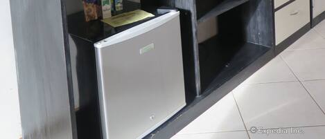 Una camera moderna e minimalista con una TV a schermo piatto su supporto nero, un piccolo frigorifero e uno scaffale con diversi oggetti, tra cui una bottiglia e una scatola.