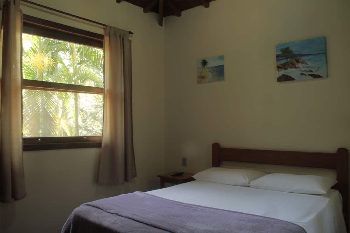 Une chambre à coucher comprenant un lit, une table de chevet, une fenêtre avec des rideaux et deux tableaux accrochés au mur.