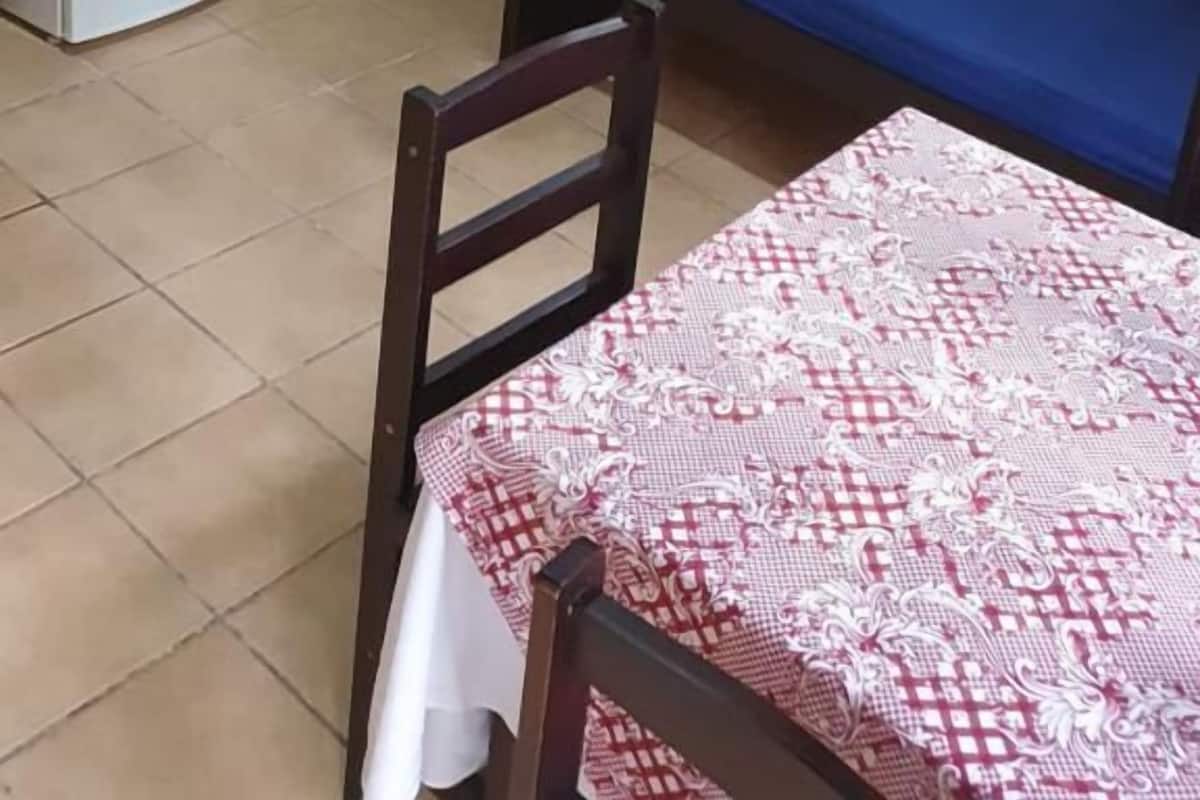 Une petite pièce avec un escalier en bois, un lit avec un matelas bleu, une table recouverte d’une nappe à motifs et un réfrigérateur blanc.