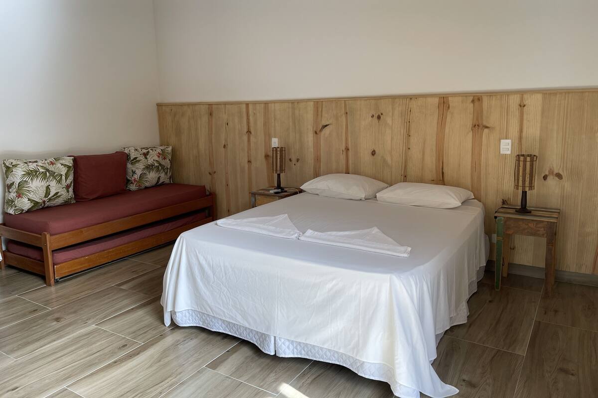 Une chambre à coucher avec une tête de lit en bois, un lit recouvert de linge de lit blanc, un canapé rouge agrémenté de coussins à motifs floraux et deux lampes de chevet.
