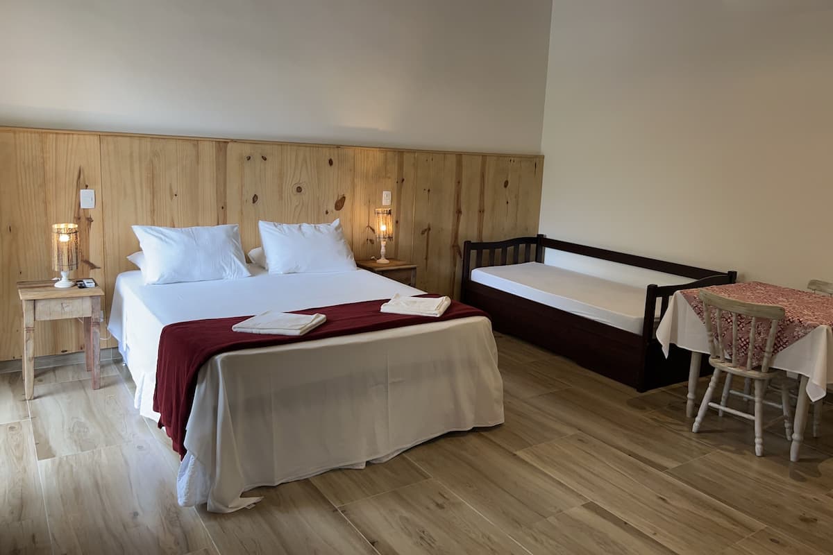 Une chambre d’hôtel avec une tête de lit en bois, un lit recouvert de draps blancs, une petite table de chevet avec une lampe et une chaise en bois placée devant une petite table recouverte d’une nappe.
