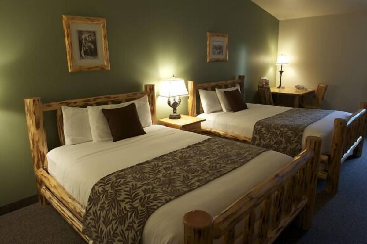 Quarto com duas camas de madeira, cada uma acompanhada de uma mesa de cabeceira e uma luminária. Há quadros nas paredes.