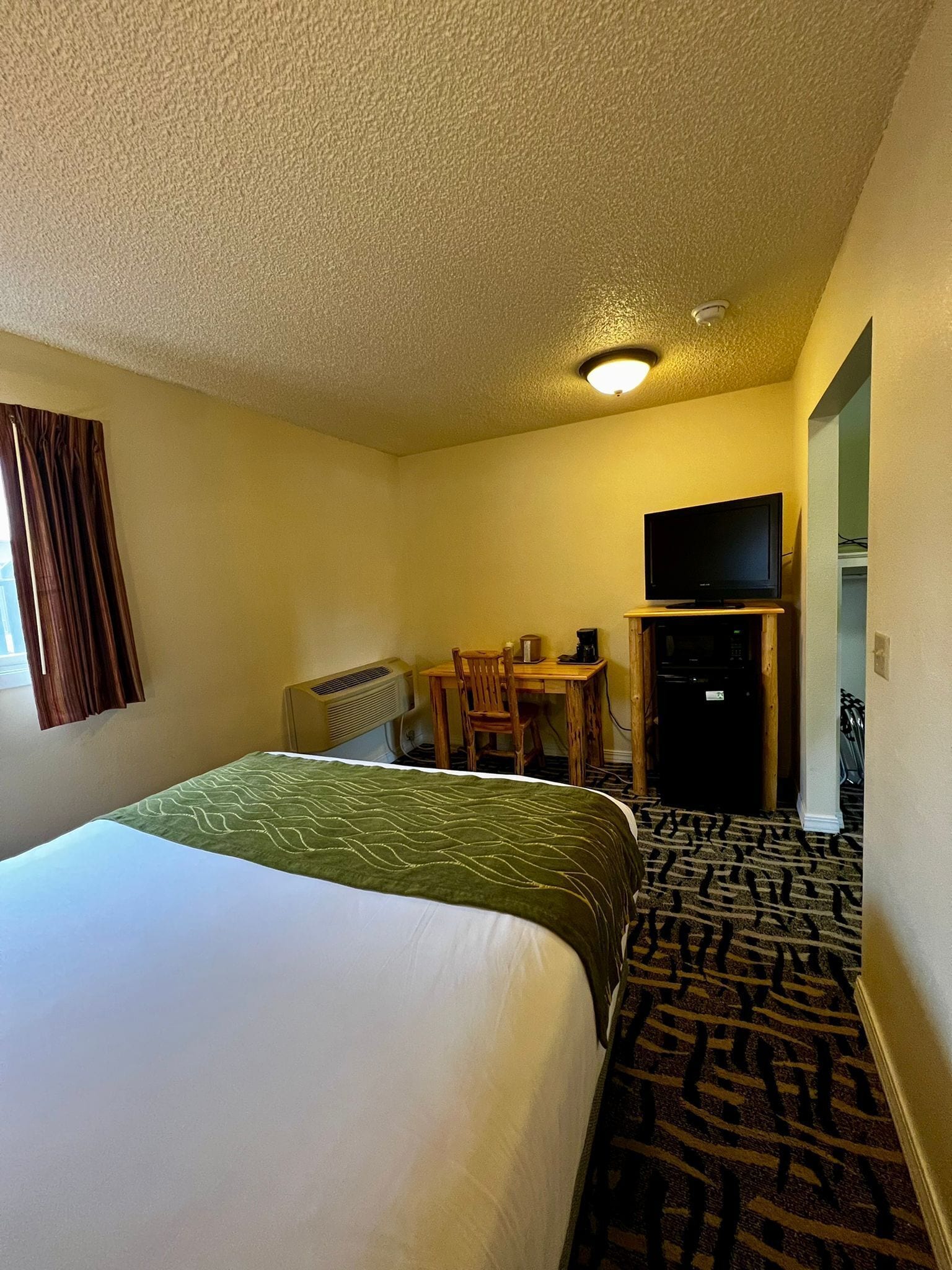 A hotel room with a large bed, a desk with a chair, a television, and a patterned carpet.