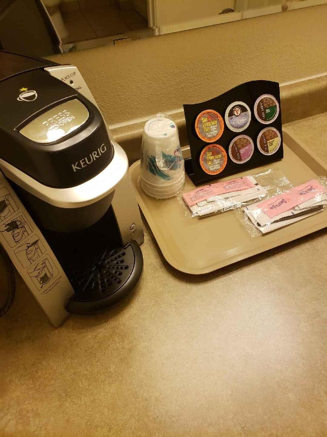 Una cafetera Keurig con un portacapsulas, una taza reutilizable y una bandeja con cápsulas de café y una bolsa de café.