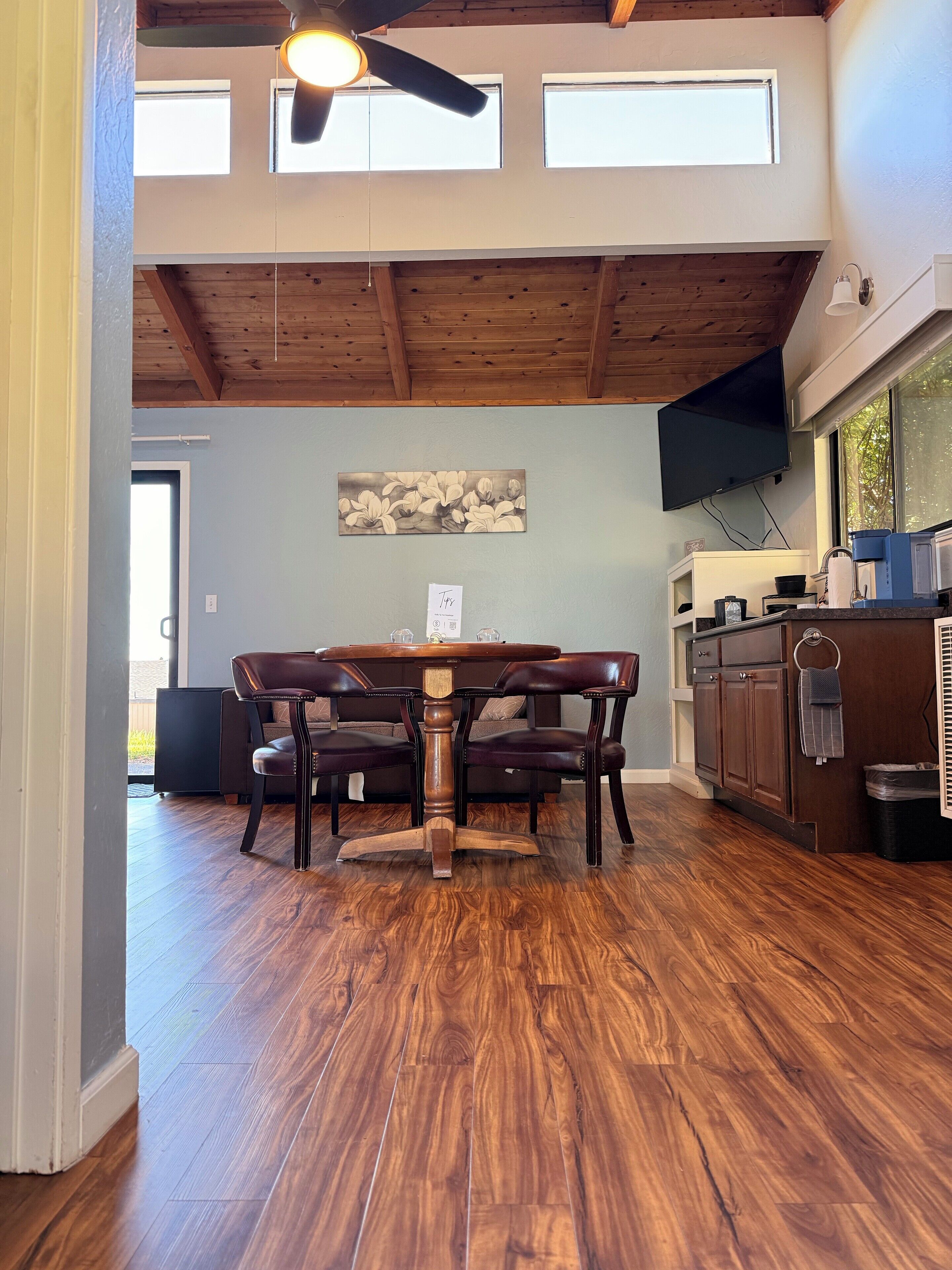 A dining area with a wooden table and chairs, a ceiling fan, and a kitchen with cabinets and a window.