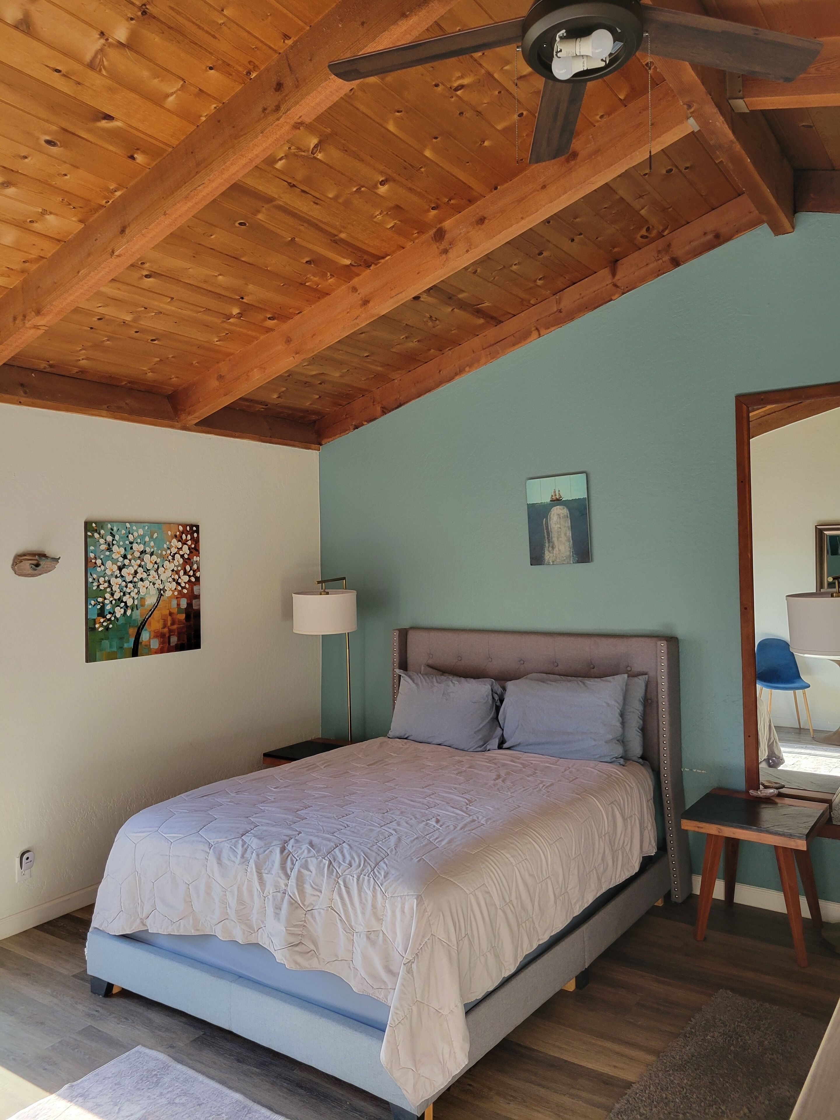 A bedroom with a wooden ceiling, a bed with a headboard, a nightstand, a lamp, and artwork on the walls.
