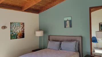 A bedroom with a wooden ceiling, a bed with a headboard, a nightstand, a lamp, and artwork on the walls.
