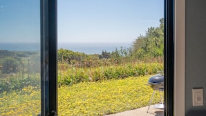 A view from a room with a large glass door, overlooking a garden with yellow flowers and a coastal landscape.