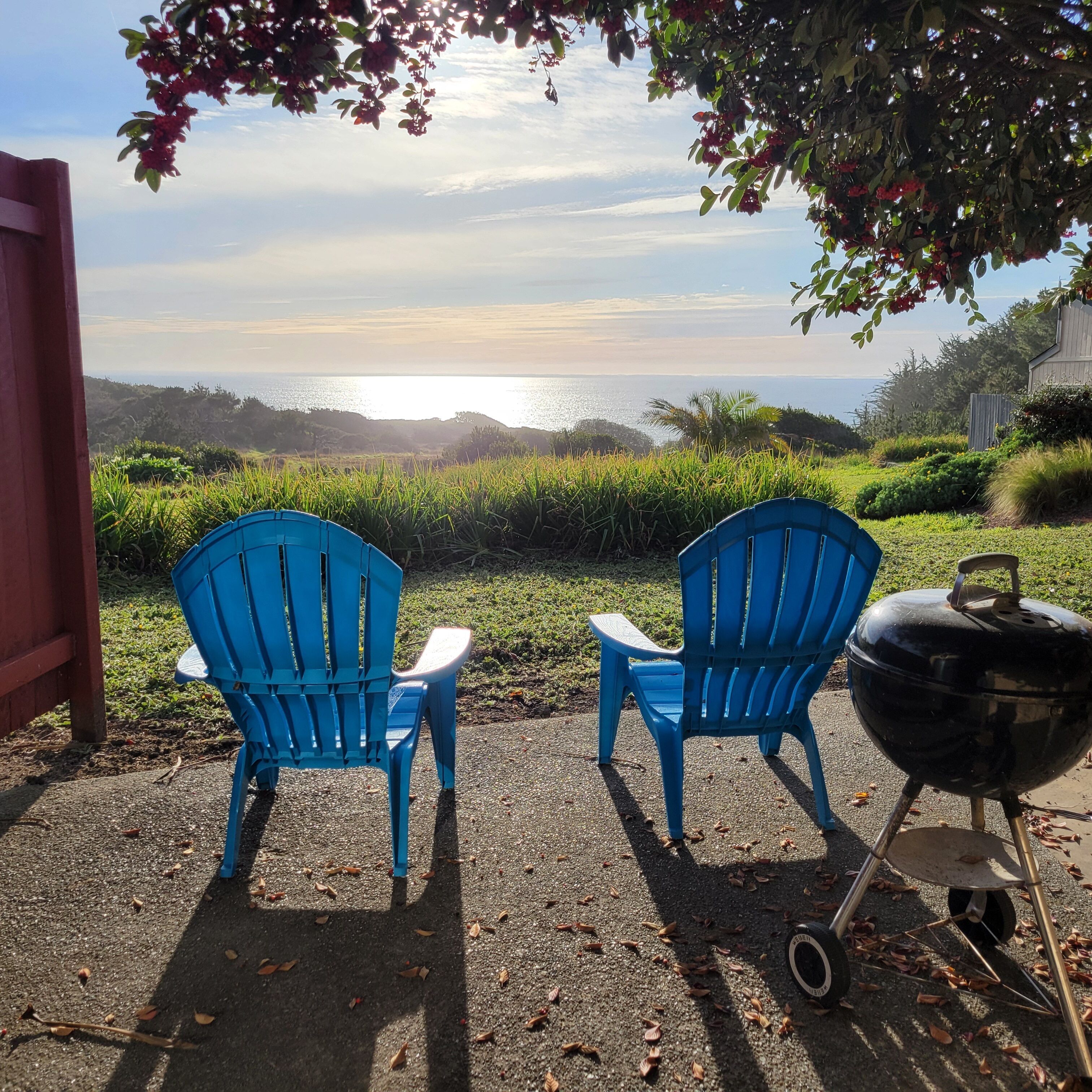 Two blue Adirondack chairs facing a scenic view of the ocean with a barbecue grill nearby.