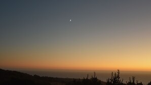 A clear sky with a crescent moon and a sunset over a coastal landscape.