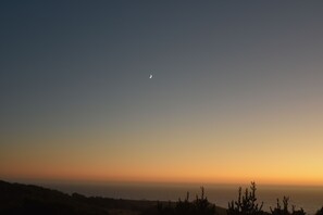 A clear sky with a crescent moon and a sunset over a coastal landscape.