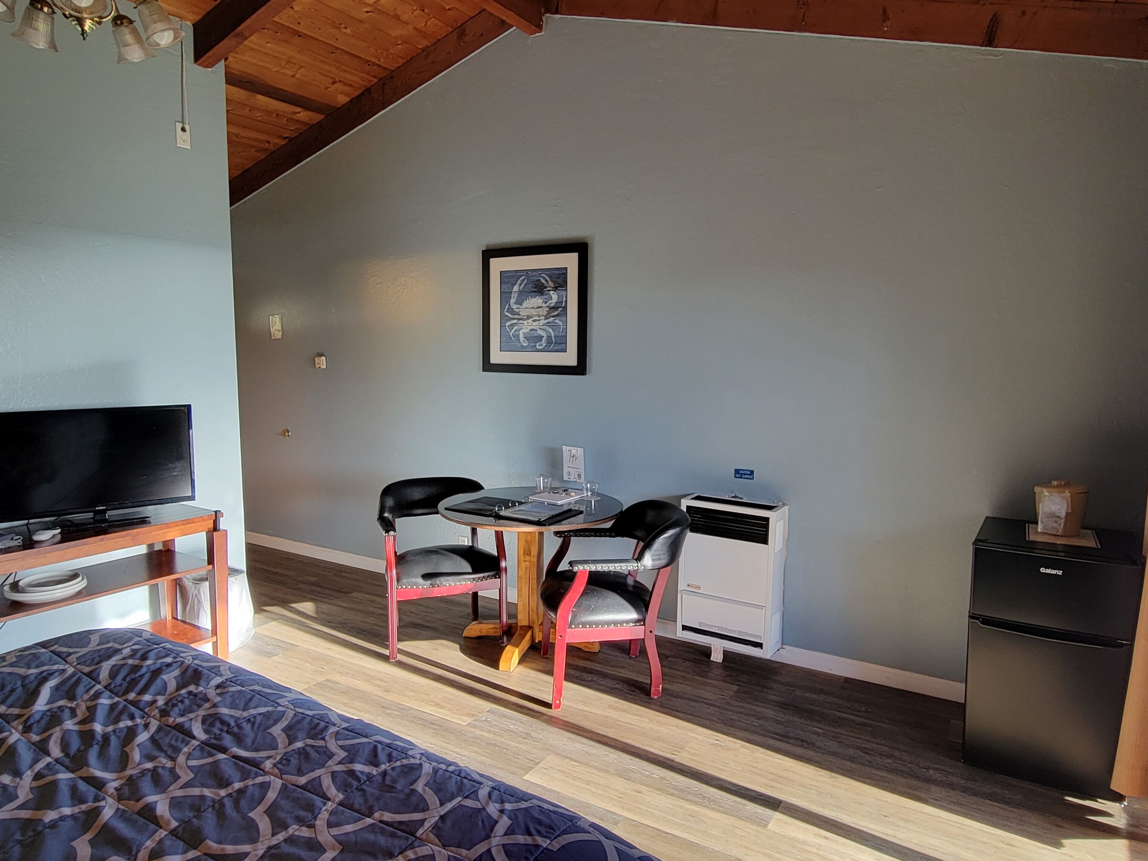 Een slaapkamer met een bed, een televisie op een houten standaard, een bureau met twee stoelen, een kleine tafel en een koelkast.