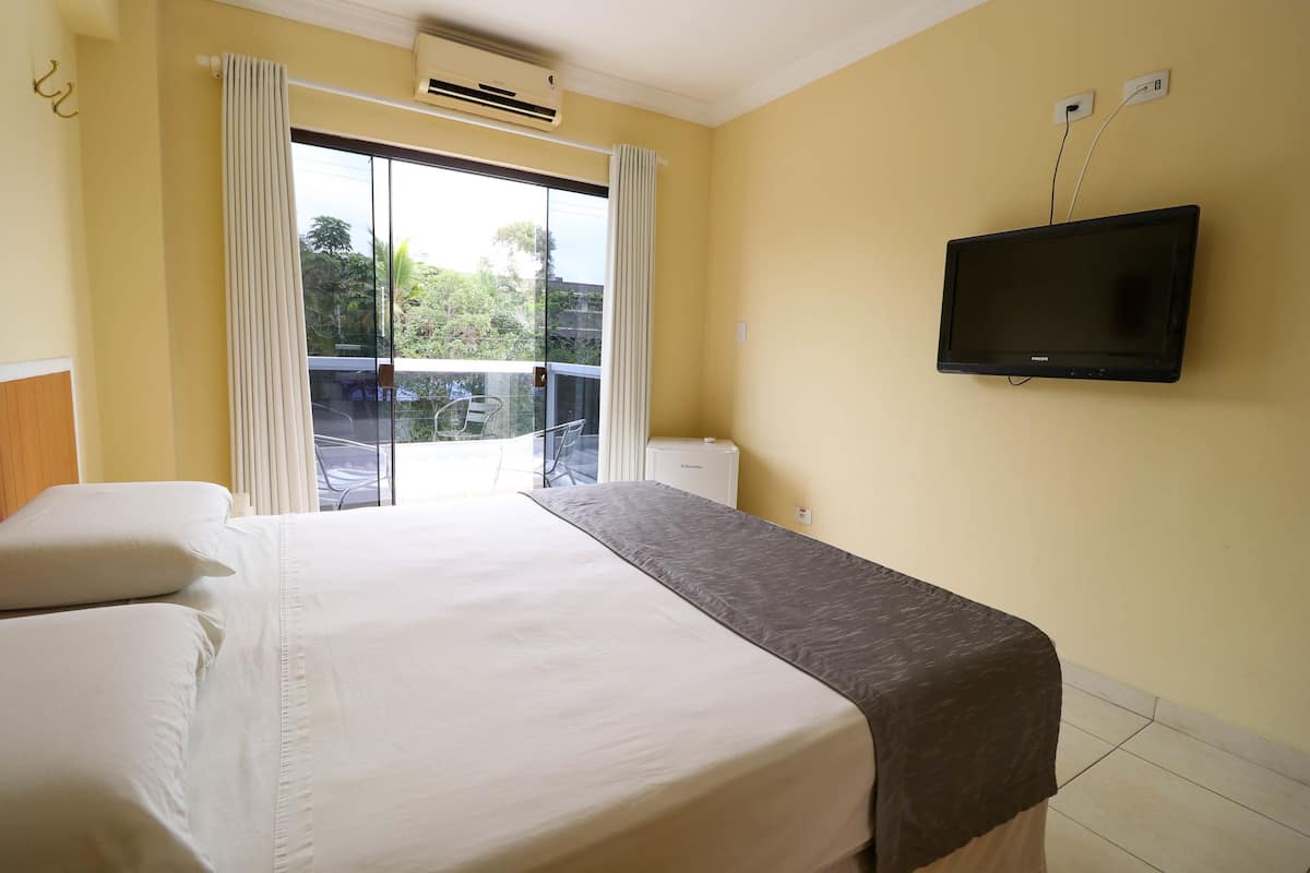 Habitación de hotel con cama, televisor montado en la pared y puerta corrediza de vidrio que da a un espacio exterior.