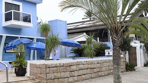 Hotel met een blauwe gevel, nummer 60, met een overdekte ingang, parasols, een palmboom en een stenen muur.