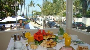 Een ontbijttafel met verschillende soorten eten, een waterkoker en uitzicht op een straat met geparkeerde auto's en palmbomen.