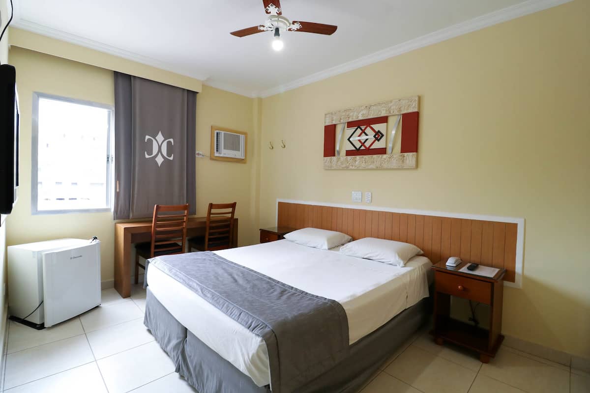 Habitación de hotel con cama, escritorio, silla, mesita de noche, ventilador de techo y ventana con cortinas.