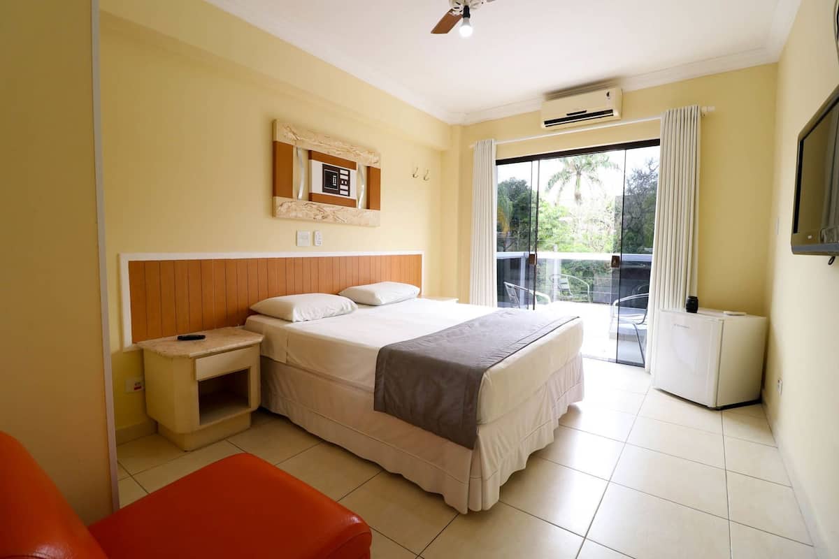 Un dormitorio con cama, mesita de noche, silla, televisor y balcón con vistas.