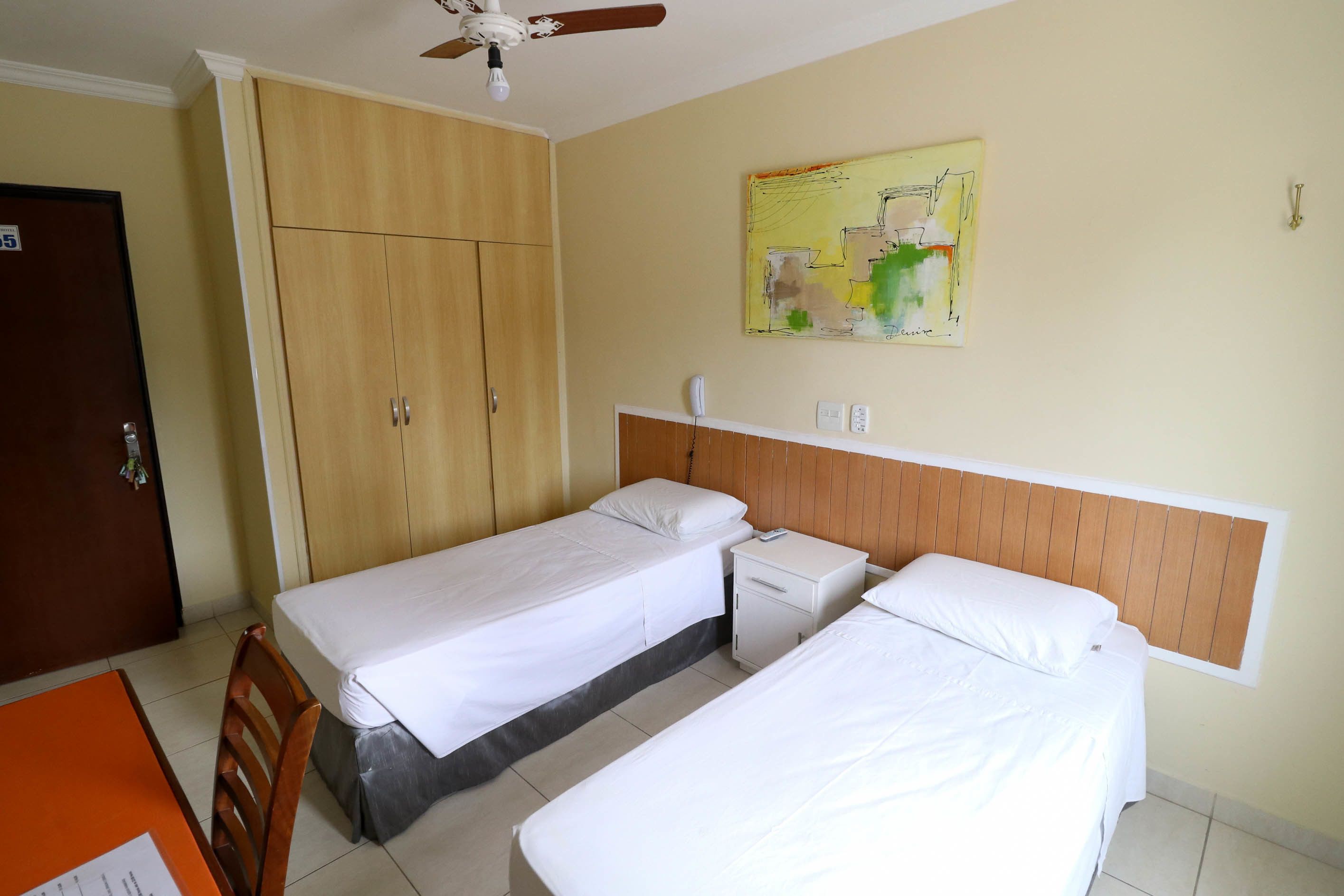 A hotel room with two single beds, a wooden wardrobe, a small bedside table with a lamp, and a ceiling fan.