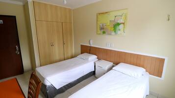 A hotel room with two single beds, a wooden wardrobe, a small bedside table with a lamp, and a ceiling fan.
