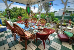 A covered outdoor dining area with wicker chairs and a glass table, surrounded by lush greenery and a view of a residential area. - Hale Kawehi B&B Guesthouse (Hilo)