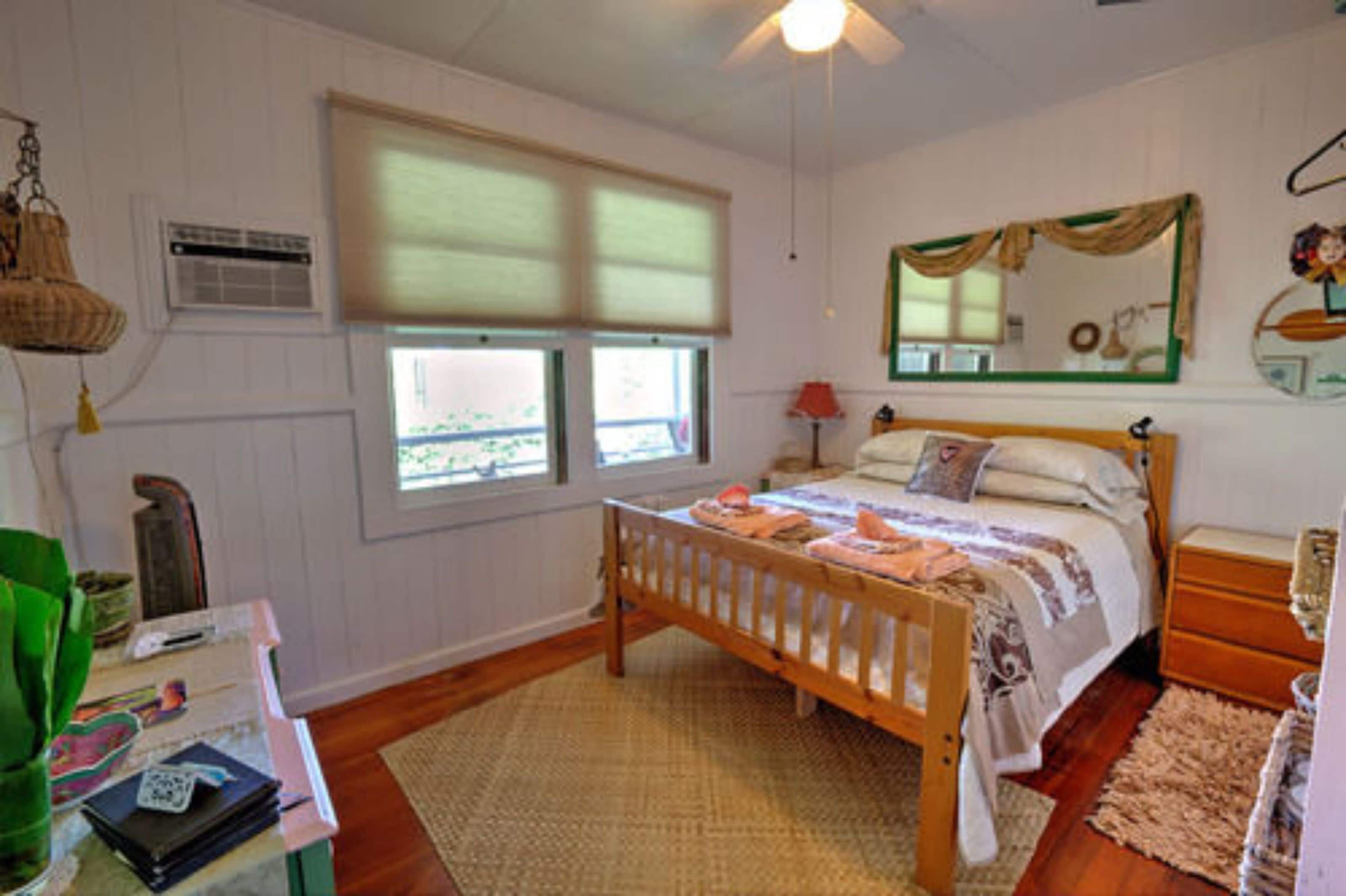 A bedroom with a wooden bed, a bedside table, a nightstand, a mirror, and a window with blinds.