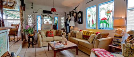 A cozy living room with a sofa, coffee table, and a ceiling fan. There are decorative items and a window with artwork.
