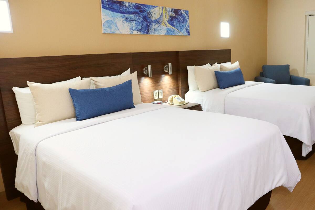 Habitación de hotel con dos camas, cada una con ropa blanca y almohadas azules, una mesita de noche con teléfono, y una obra de arte colgada en la pared.