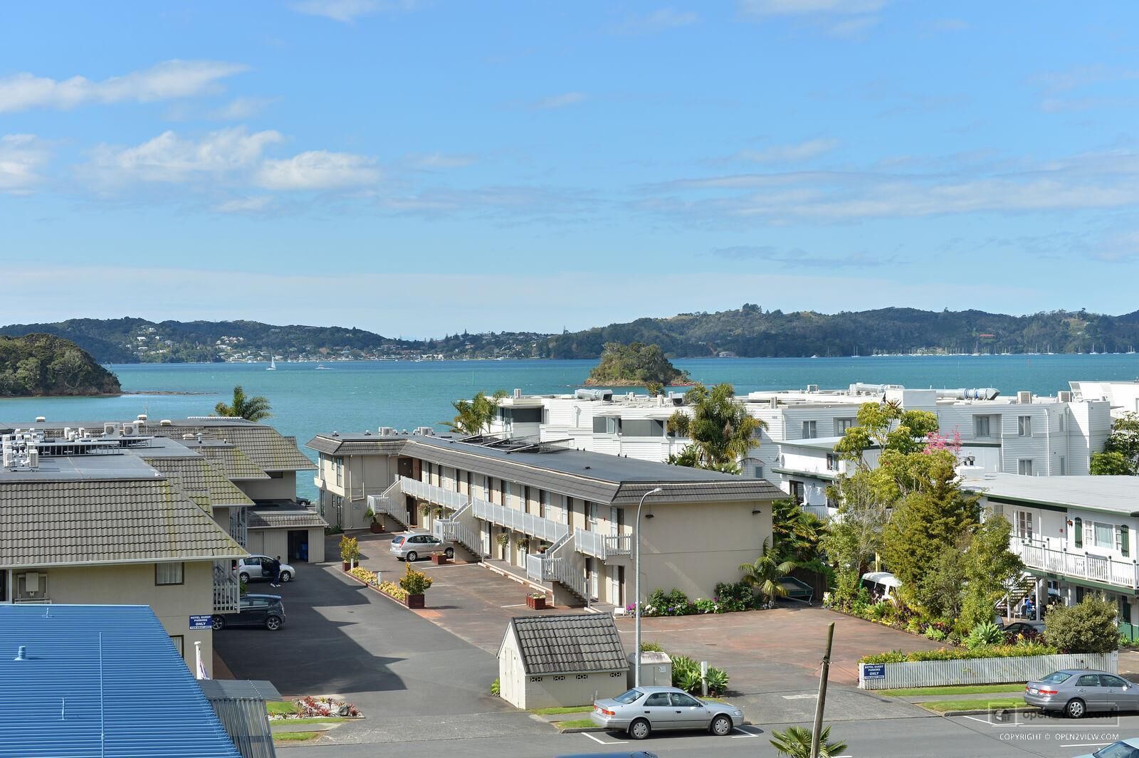 A coastal residential area with multiple-story buildings, a parking lot, and a view of the sea and distant hills.