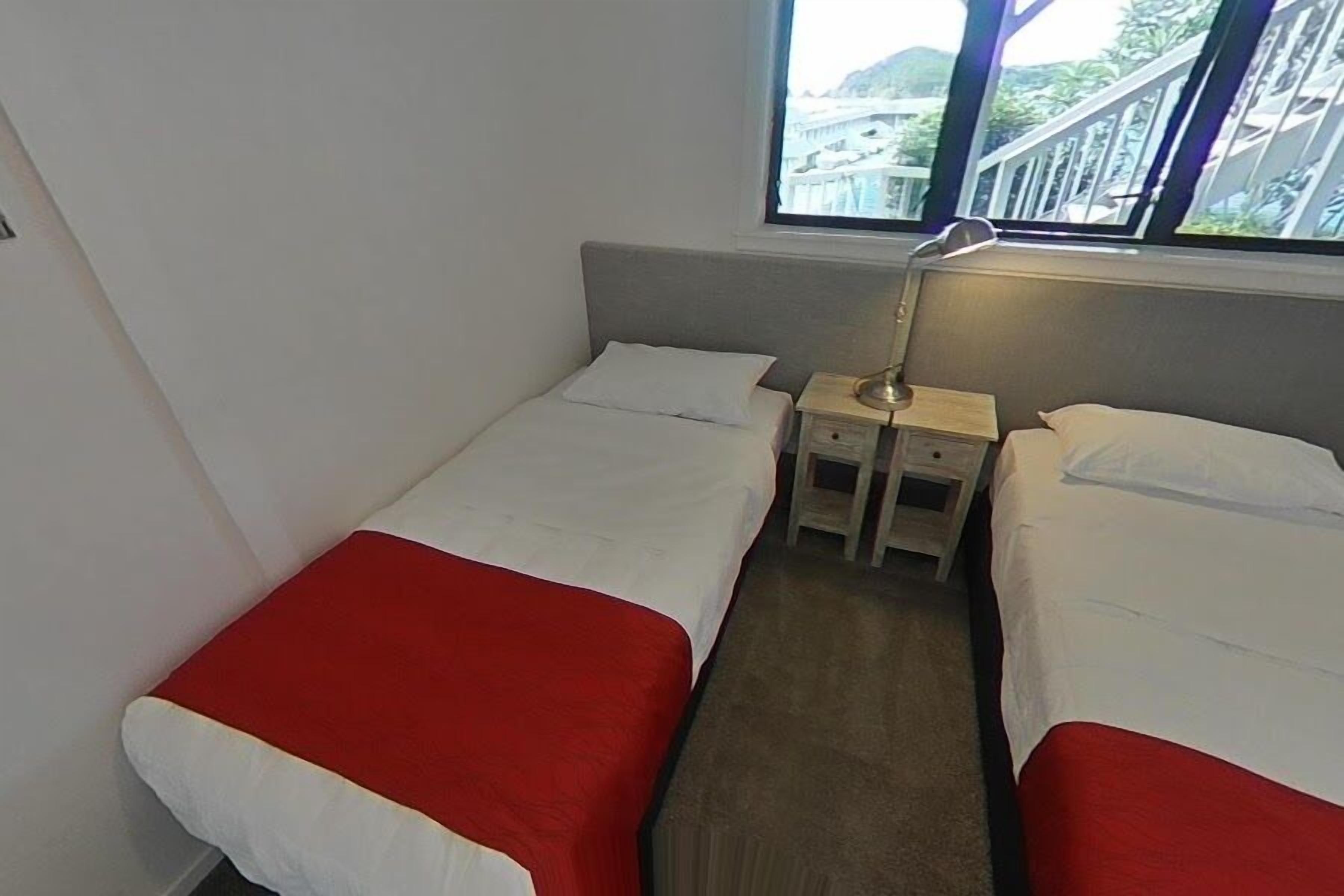 Habitación de hotel pequeña con dos camas individuales, una mesita de noche, una lámpara y ventana con vistas a un puente y vegetación.