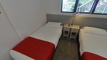 Habitación de hotel pequeña con dos camas individuales, una mesita de noche, una lámpara y ventana con vistas a un puente y vegetación.