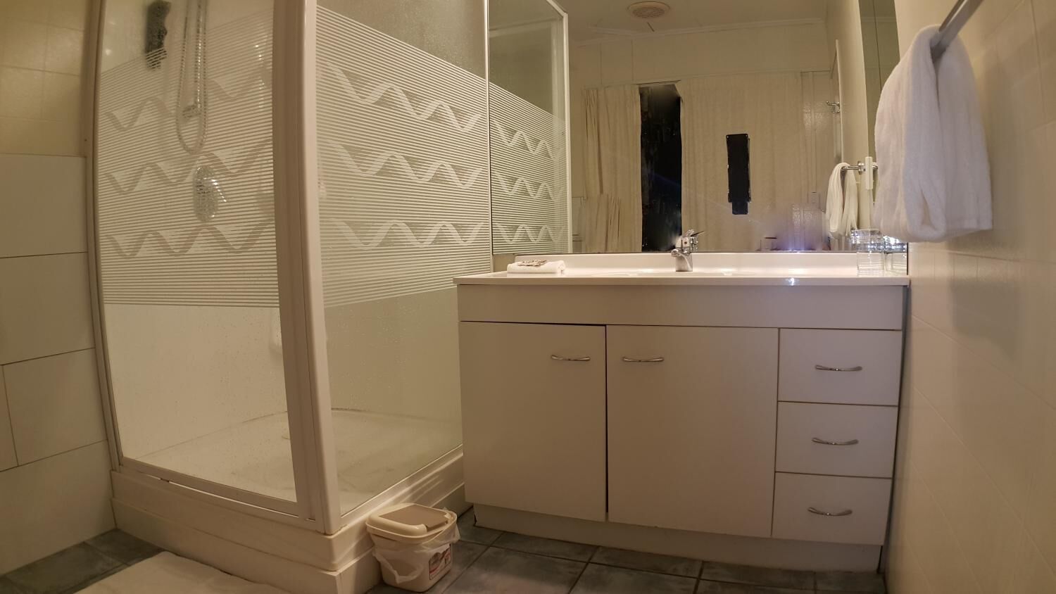 A bathroom with a glass shower enclosure, a white vanity with a mirror, and a towel hanging on the wall.