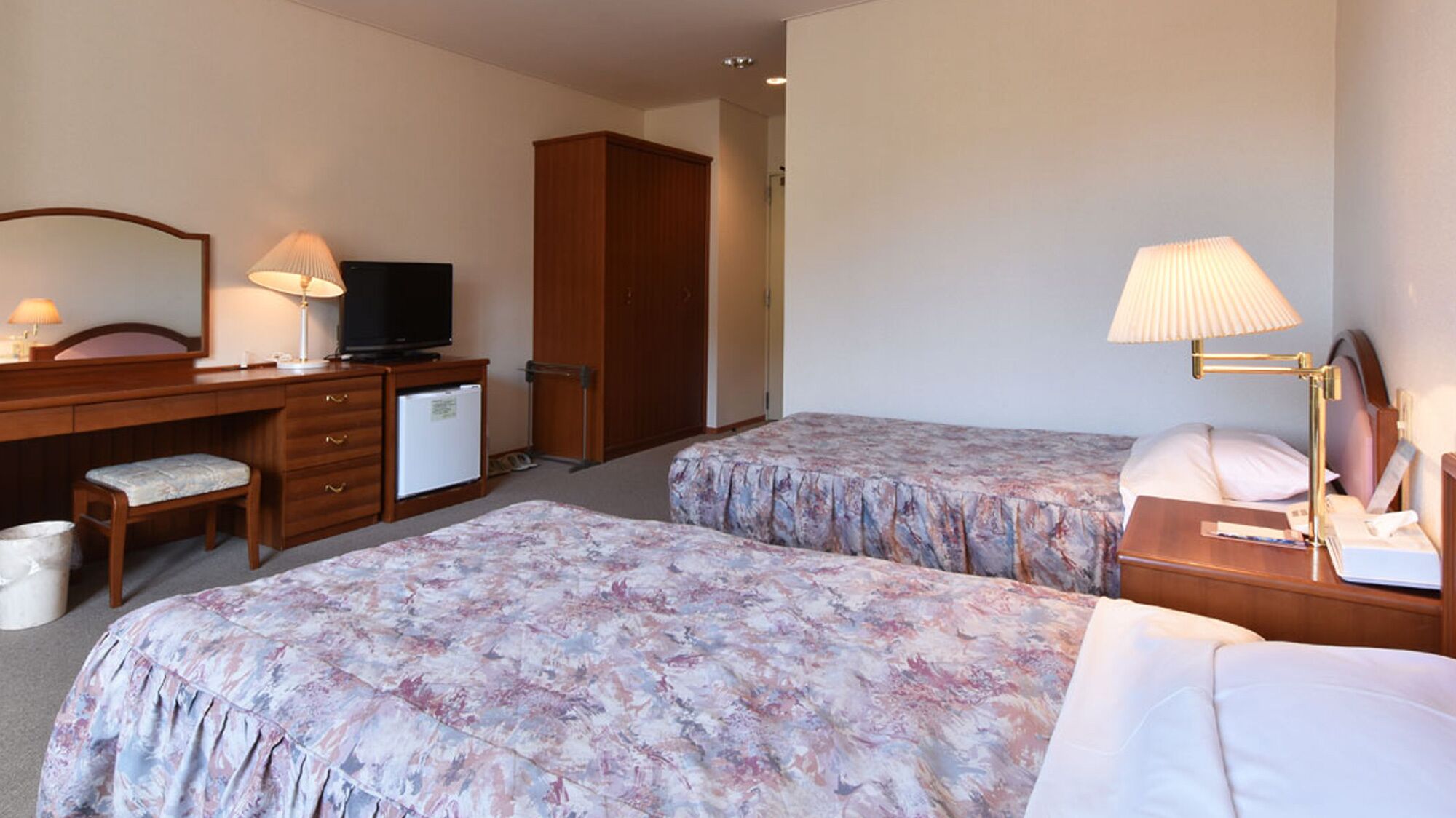 A hotel room with two beds, a desk with a television, a small refrigerator, a chair, and a nightstand with a lamp.