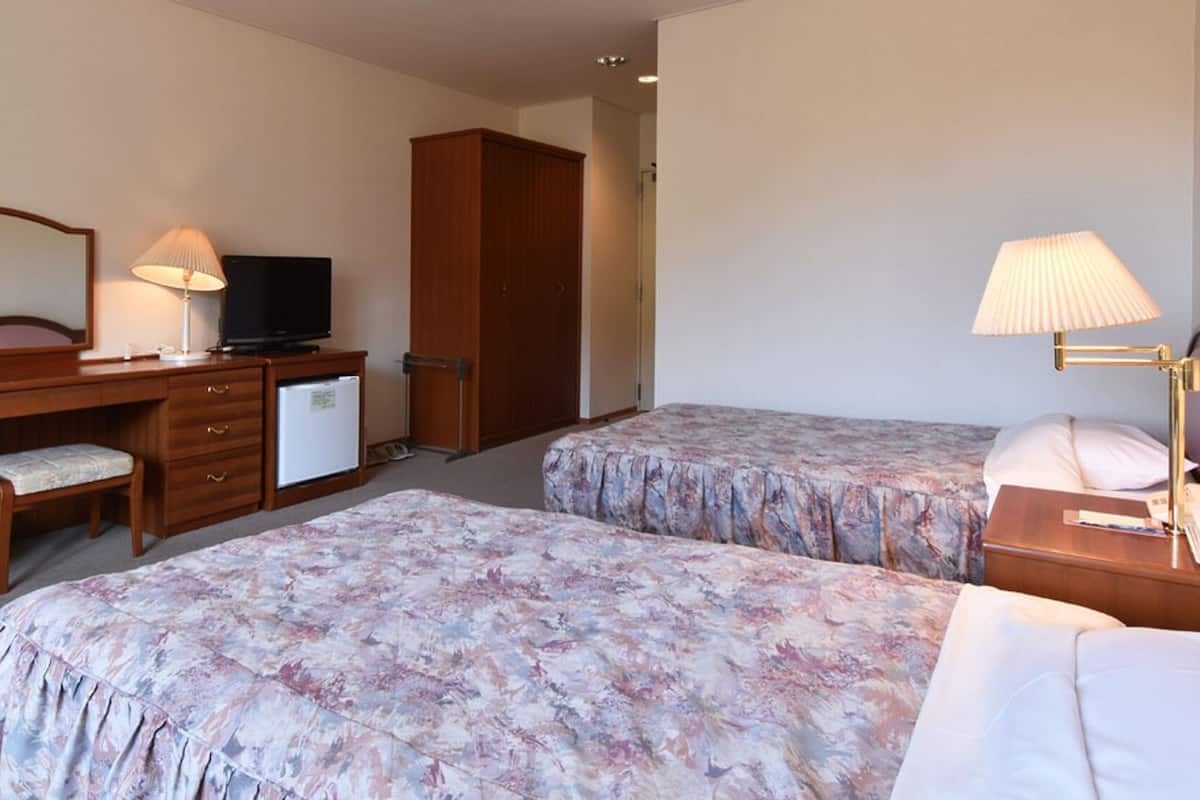 Hotellihuone, jossa on kaksi sänkyä, työpöytä, jossa on televisio, pieni jääkaappi, tuoli ja yöpöytä, jossa on lamppu.