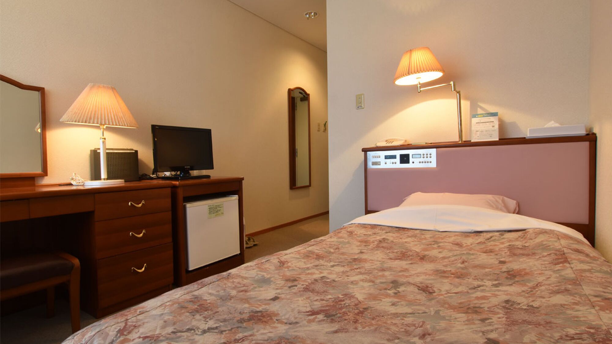 A hotel room with a bed, a desk with a computer, a lamp, a mirror, and a thermostat on the wall.