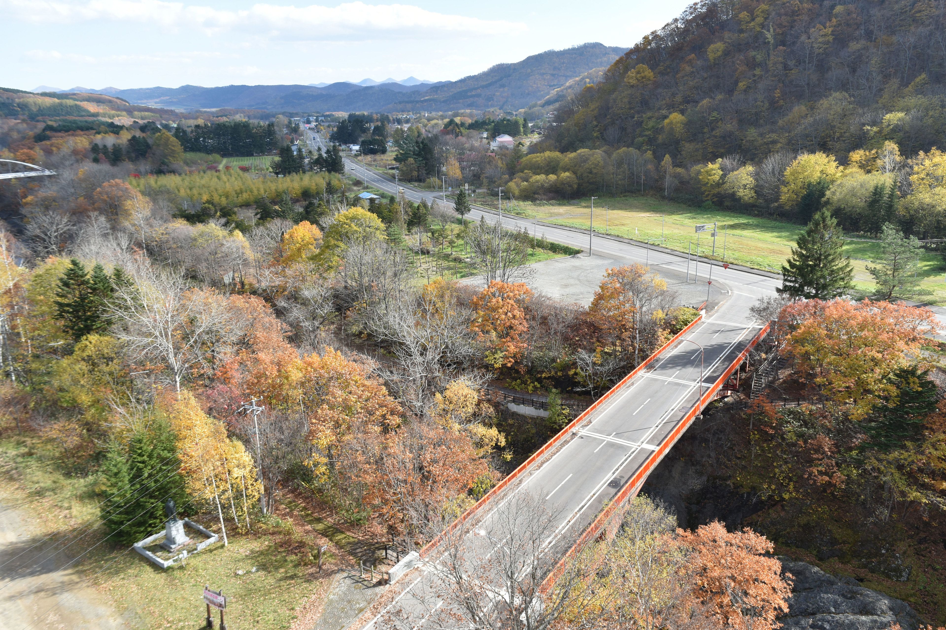 A bridge over a road with autumn foliage, surrounded by hills and a distant town.