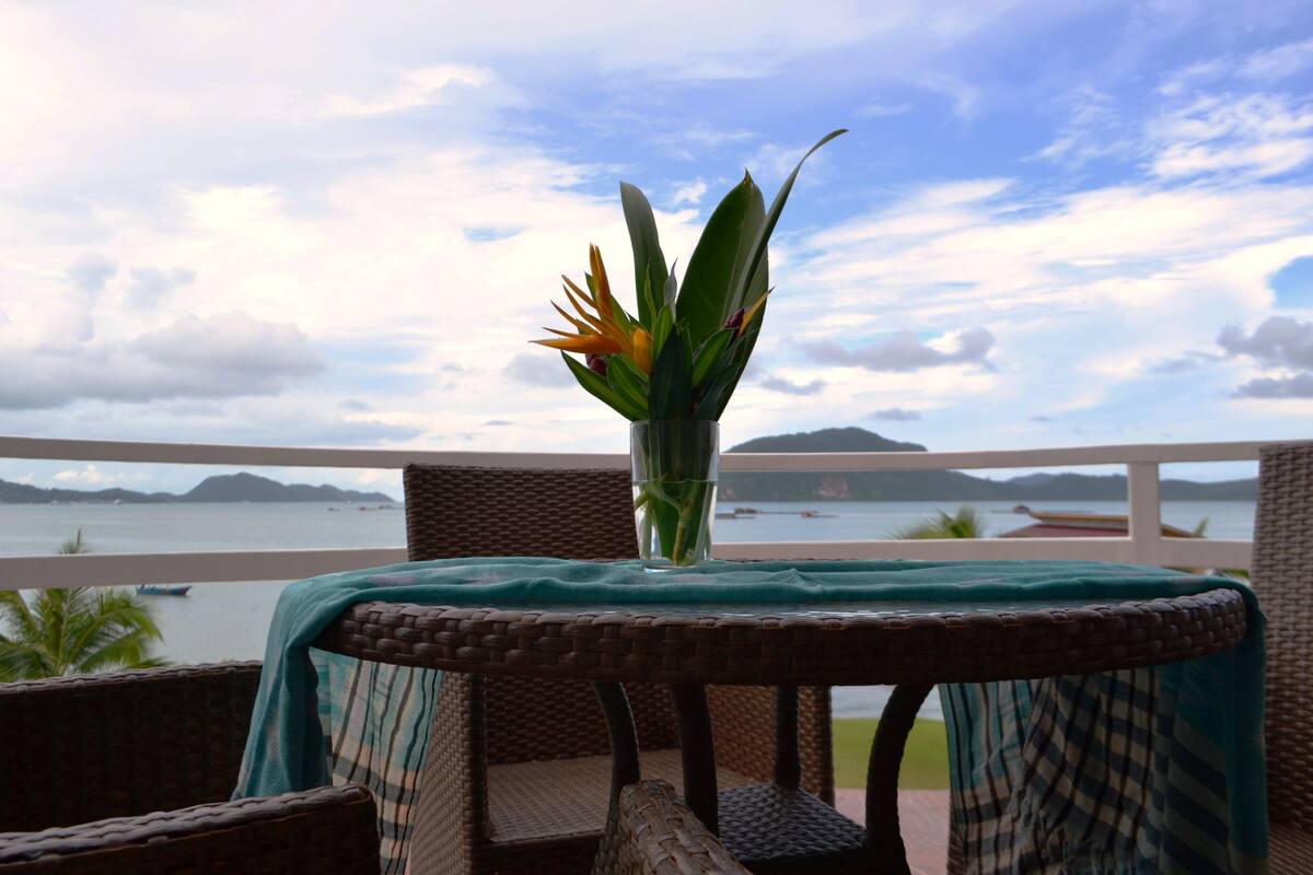 A wicker chair with a table covered by a green cloth, a vase with a plant, and a view of the sea and mountains.