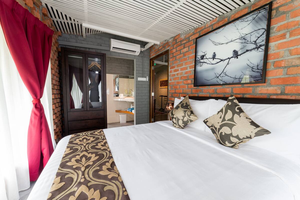 A hotel room with a large bed, a red curtain, a brick wall, and a framed picture of birds on a branch.