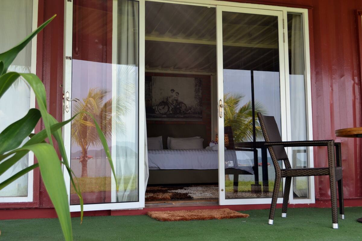 A room with a bed, a chair, and a small table. There is a sliding glass door leading to the outside.