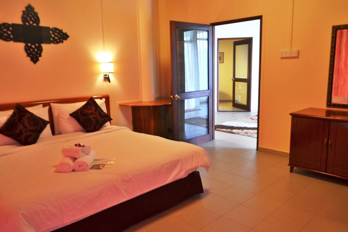 A hotel room with a bed, pillows, a nightstand, a dresser, and a door leading to a hallway.