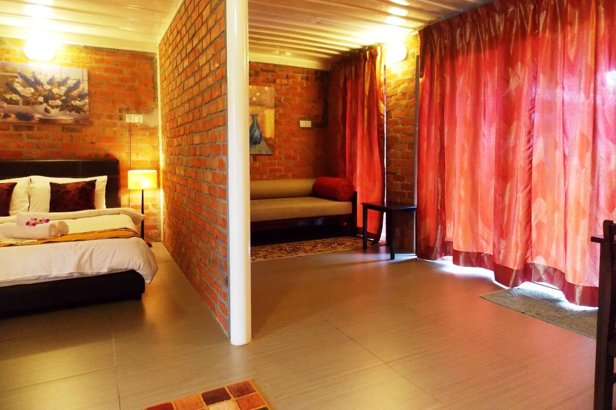 A hotel room with a brick wall, a bed with white linens, a sofa, and red curtains.