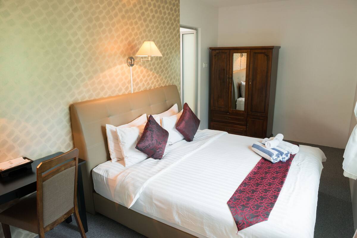 A neatly made bed with a red and white patterned bed runner, a wooden nightstand with a lamp, and a wooden wardrobe in a hotel room.