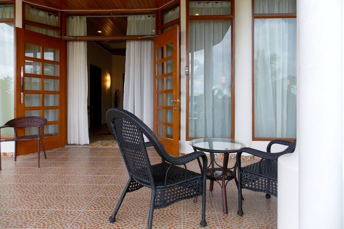 A covered outdoor seating area with wicker chairs and a table, wooden doors with glass panels, and a tiled floor.