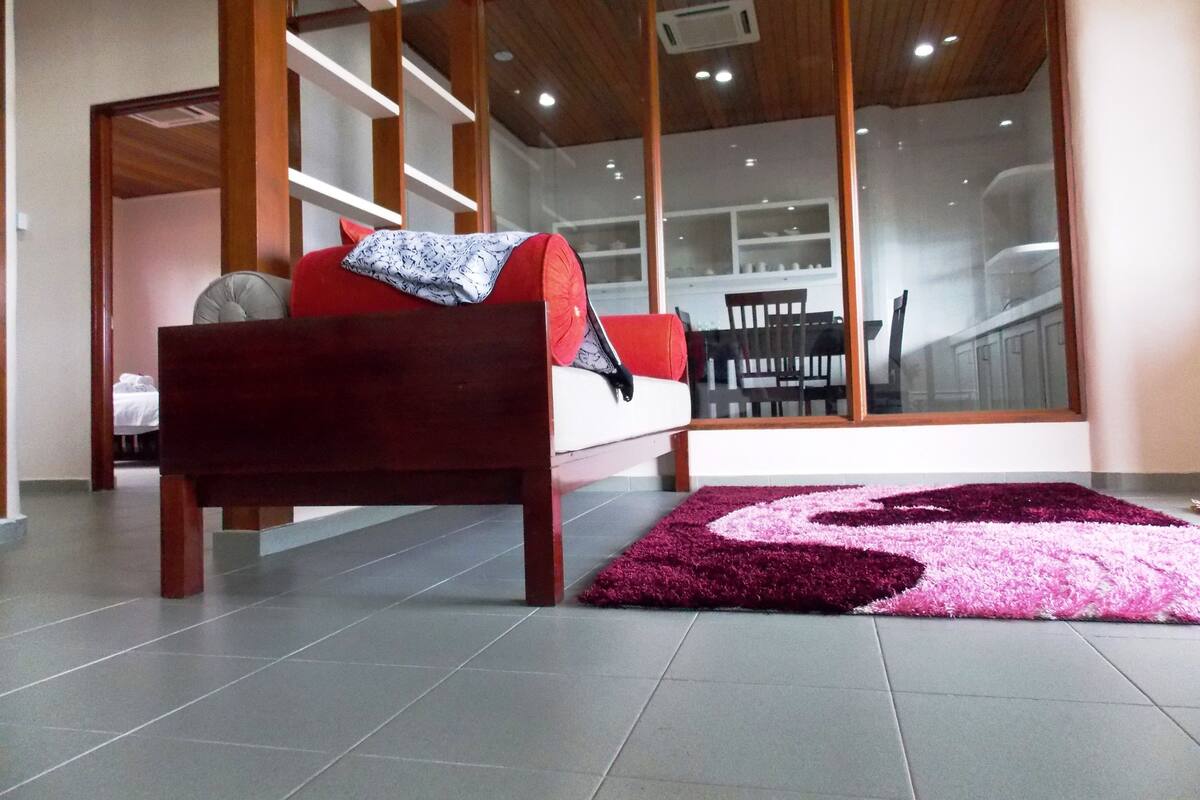 A modern bedroom with a wooden bed, a red and white sofa, and a large window offering a view of the kitchen.